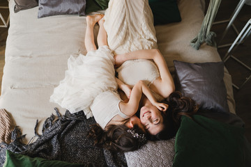 Mother and daughter hugging and cuddling in bed. Pretty little girl and beautiful woman lie together. Girls in lace dresses playing in decorated room. Family weekend, beauty day, having fun, love.