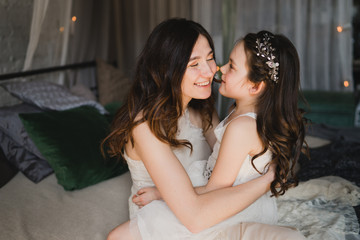 Mother and daughter hugging and cuddling in bed. Pretty little girl and beautiful woman lie together. Girls in lace dresses playing in decorated room. Family weekend, beauty day, having fun, love.