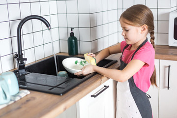 A cute little European girl washes dishes in an apron in the bright kitchen. The child helps in the kitchen to wash and wipe dishes.