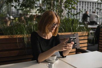 Young business woman uses phone. Good looking brunette female uses online banking on smart phone to transfer money from credit card. Girl using smart phone and chatting with business partners in cafe.