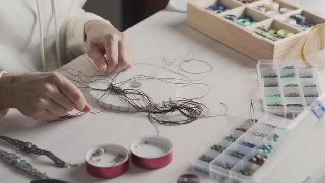 Lifestyle concept, work from home to reinvent your life: close-up of woman hands making macrame knotted jewelry with stone beads and tools on light wooden table