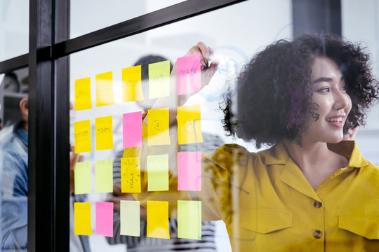 Young Creative Team Use Post It Notes To Share Idea Sticky Note On Glass Wall. And Working Design Planning And Brainstorming Concept. Business Woman And Asian Business People Meeting At Modern Office