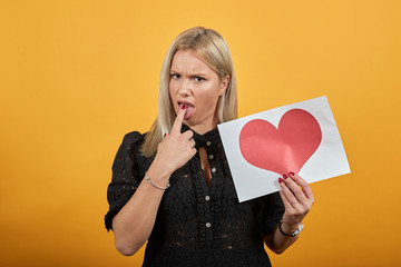 Beautiful blonde girl in black dress on yellow background holds a piece of paper with red heart and...