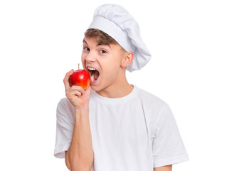 Happy teen boy in chef hat holding and eats fresh red apple, isolated on white background. Portrait of smiling child cook bites delicious fruit. Organic natural healthy food produce.