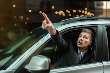 young businessman pointing with his finger from the open car window, looks amazed, big city life