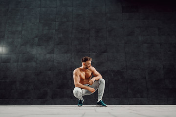 Handsome caucasian shirtless muscular blond bearded man crouching in front of dark background.