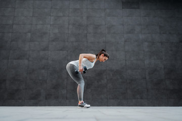 Fototapeta na wymiar Side view of fit healthy caucasian brunette in sportswear bending and doing exercises with dumbbells in hands. In background is dark wall.