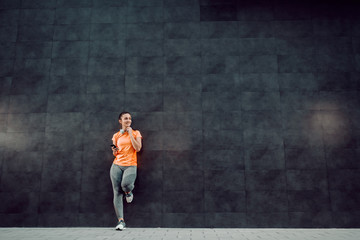 Full length of attractive caucasian sportswoman in shape leaning on dark wall, holding smart smart phone and looking away.