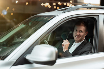 young smiling businessman looks happy while driving his car with open window, multitasking, big city life