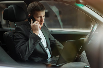 young businessman works in his car using his laptop having conversation on smartphone getting ready for important meetting with partners, multitasking concept