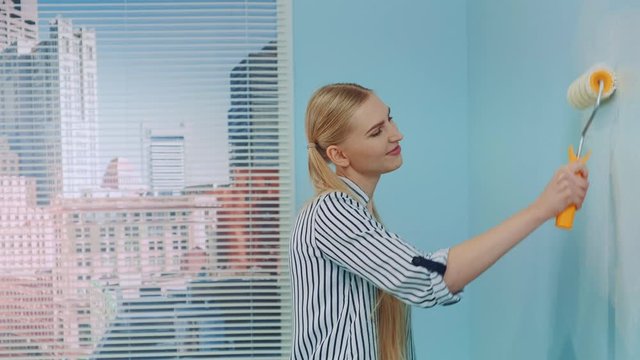 Portrait shot of a young beautiful woman painting a wall at office. She uses white paint and work with a roller. Office renovations.
