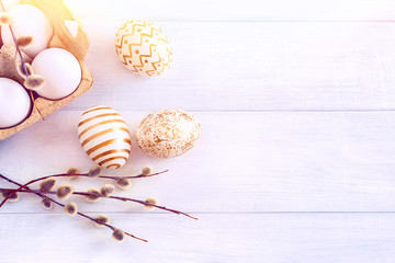 Sunny Easter morning. Unpainted and painted with gold patterns chicken eggs in a cardboard box with a willow branch on a white table with sunlight close-up. Festive background, top view, copy space.