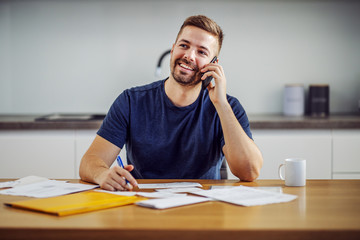 Young attractive handsome bearded smiling man calling customer service while sitting at dining table and filling in bills.