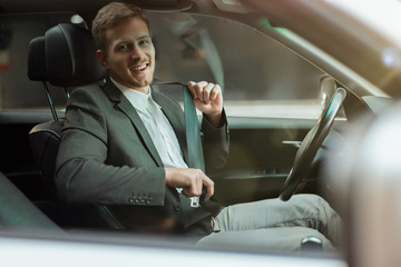 young smiling businessman sitts in his car looks happy and satisfied, adjusting his safety belt...