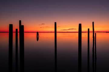 A long exposure photo of pylons in the sea at sunset with Venice in the background