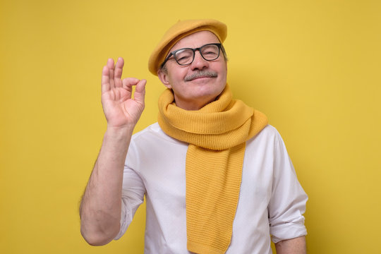Hispanic senior man in glasses and yellow beret and scarf looking with a smile at camera showing ok sign. Studio shot. Positive facial human emotion.