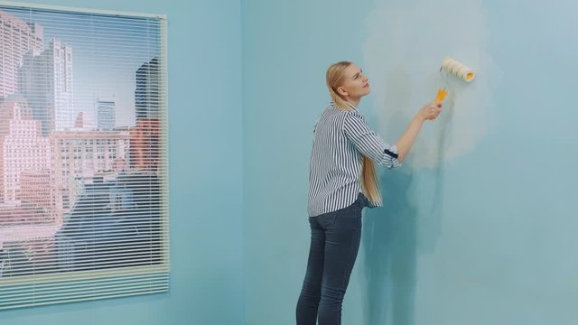 Beautiful woman renovating her new apartment. She uses white paint and work with a roller. In the background there are skyscrapers at day.