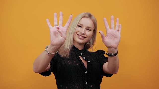 Beautiful blonde girl in black dress on yellow background happy woman shows nine fingers and smiles