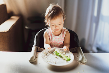 A nine-month-old smiling baby girl sits at a white table in a highchair and eats herself with a spoon from a bowl. Dark background. Healthy eating for kids. Child's nutrition.