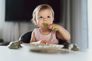 A nine-month-old smiling baby girl sits at a white table in a highchair and eats herself with a...
