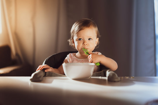 Nine-month-old smiling baby girl in pink bandage on her head sits at white table in highchair and eats herself with spoon from bowl. Blurred background. Healthy eating for kids. Child's nutrition.