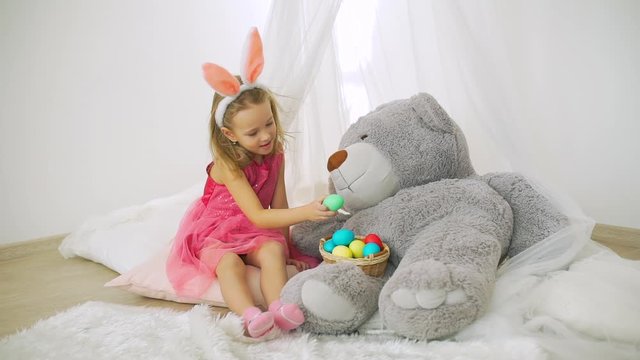 Cute Little Girl Sitting on the Floor in Nursery and Feeding Big Toy Bear Colorful Eggs. Easter Holiday Celebration and People Concept