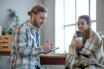 Young man standing writing, girl sitting drinking coffee.