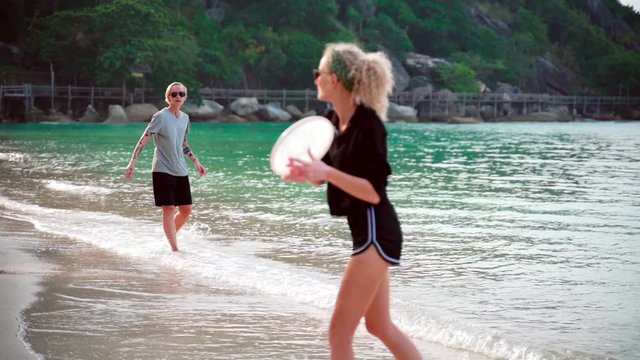 Slow-motion footage of two young women throwing a frisbee at the tropical beach on a sunny day.