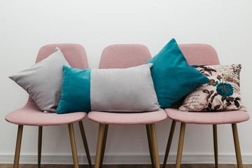 Different pillows on three pink chairs in room, against white wall background