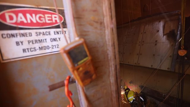 Footage of danger confined space sign entry by permit only gas test atmosphere detector monitoring rope access welder wearing safety harness abseiling conducting hot work a oxygen cutting steel plate 