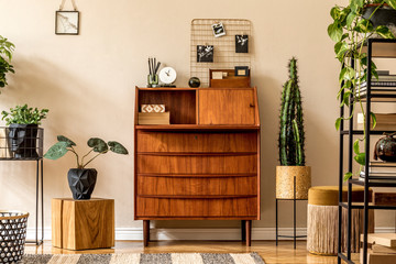 Retro interior design of living room with wooden vintage bureau, shelf, gold pouf, cube, plants, cacti, books and elegant personal accessories. Stylish vintage home decor. Beige wall. Template. 