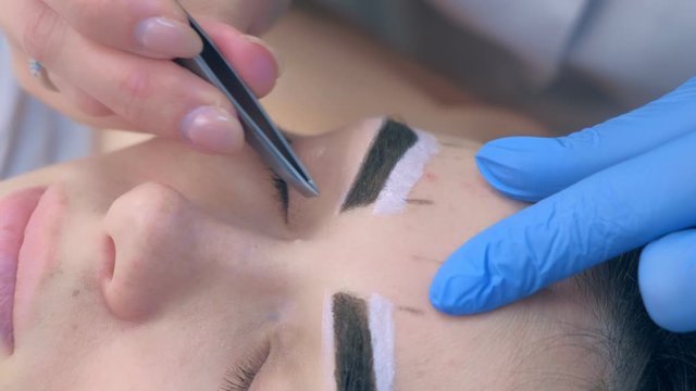 Professional beautician plucking eyebrows with tweezers to woman in beauty salon during tint eyebrow procedure, closeup view. Girl lying with closed eyes and brown natural henna on brows.