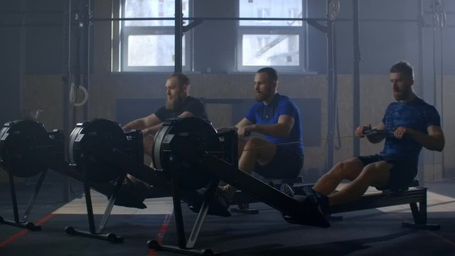 Males using on rowing machines slow motion. Sportsman exercising on row machine in gym. Fit young man exercising using rowing machine at health club. Team training.
