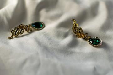 Metal gold earrings with green, emerald stones, white and yellow rhinestones. On a white fabric flattened background. Brilliant and festive decoration