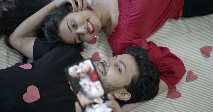 Attractive and Indian couple in red love lying down facing each other take selfie together for their pre-wedding portraits on valentine's day for their engagement fantasy slow-motion 60fps handheld 