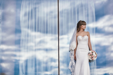 gorgeous luxury stylish bride in a white dress with a bouquet in hands near the panoramic window on the cloudy sky background.