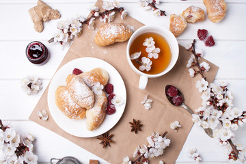 Spring card with a white cup of tea, spring branches, tasty croissants and jam on a white wooden background in flat lay style
