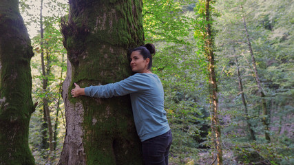 A brunette woman in the forest embraces a large tree.