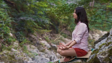 A woman with closed eyes in the Lotus position sits on a rock in the middle of the forest and meditates.