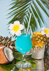 Exotic tropical Blue Curacao cocktail drink in a glass with Plumeria frangipani flower, palm leaf, fresh coconut and pineapple on the background.