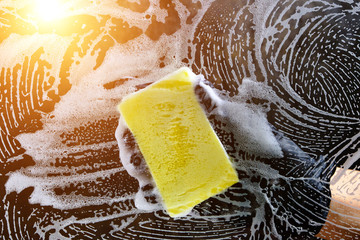 Hand in a black glove washes the glass with a foam sponge with foam on a dark background with light.