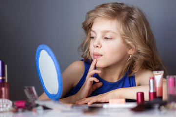 charming child, little beauty, cute girl of 7-8 years old with beautiful blond curls, make-up, mom&rsquo;s makeup at the table, gray background