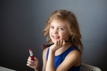 charming child, little beauty, cute girl of 7-8 years old with beautiful blond curls, make-up, mom’s makeup at the table, gray background