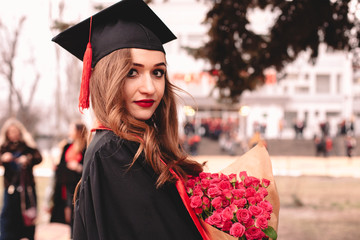 Portrait of young female graduate student in mortarboard holding bouquet of flowers while standing...