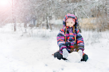 Fototapeta na wymiar Little beautiful girl sits in the snow and holds a snow globe in her hands. In colored overalls and gloves.