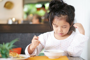 Asian child cute or kid girl enjoy eating with drink cereals soy milk or scooping and croissant bread for breakfast and healthy food in morning with holding spoon at home or restaurant and coffee shop