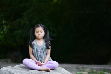 Asian child cute or kid girl close eye mindfulness meditating with breath and sit on stone or rock for peace and relax or yoga on nature green garden and tree at temple park or meditate church outdoor - obrazy, fototapety, plakaty