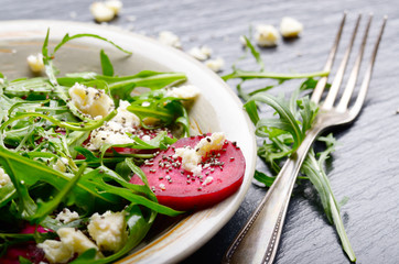 Clay dish with beetroot arugula and feta cheese salad on slate stone tray closeup view. Fork aside