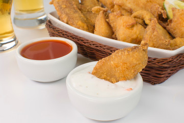 One fried tilapia strip in a bowl with tartar sauce close with a platter of fried tilapias and beer in white background