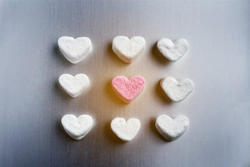Fototapeta na wymiar pink marshmallow heart lies among white sugar marshmallows square shaped, with sun flare on gray aluminum background